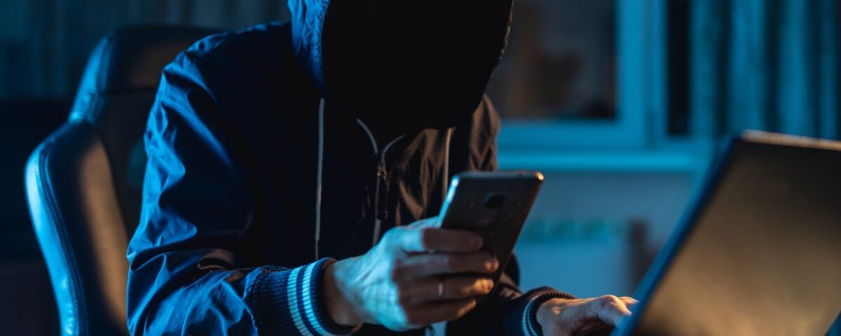 A person in a hoodie with their face in shadow sitting in front of a laptop while holding a smartphone.