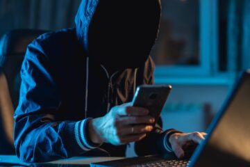 A person in a hoodie with their face in shadow sitting in front of a laptop while holding a smartphone.