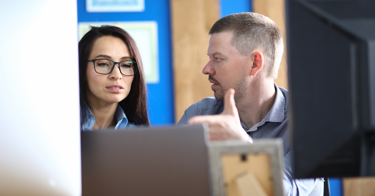 A man turns his head to speak to a woman wearing glasses sitting next to him. The backs of computer monitors frame their faces.