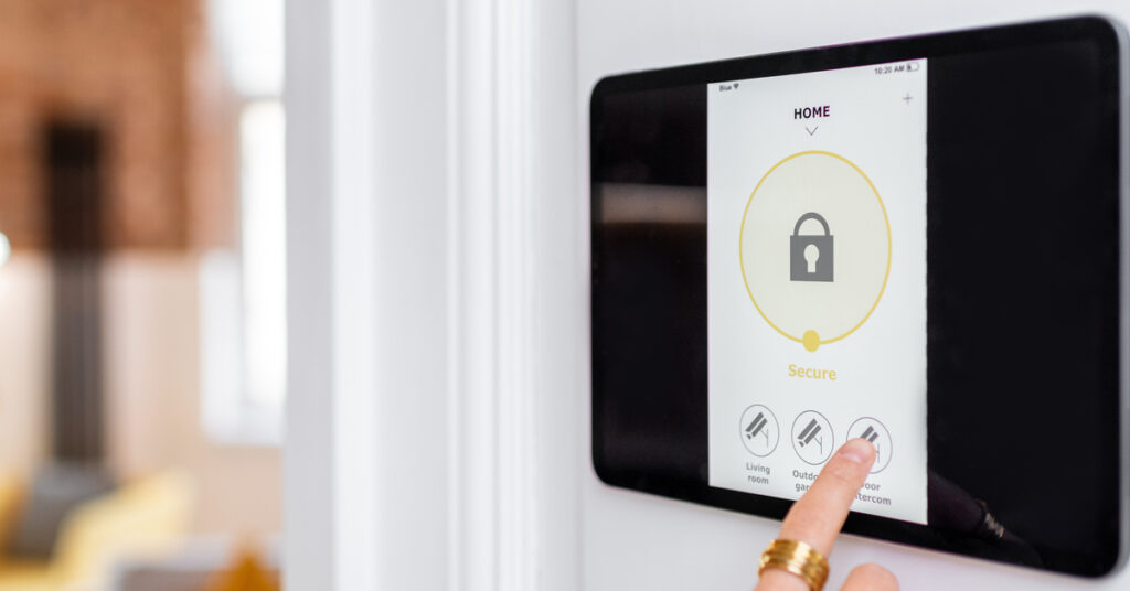 A touch-screen alarm system on a white wall. A finger with a gold ring is reaching to tap the screen.