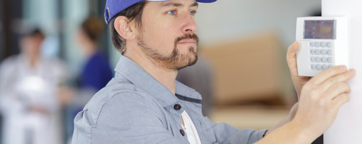 A man with a beard in a blue hat and button-up shirt is holding a white alarm console up to a wall.
