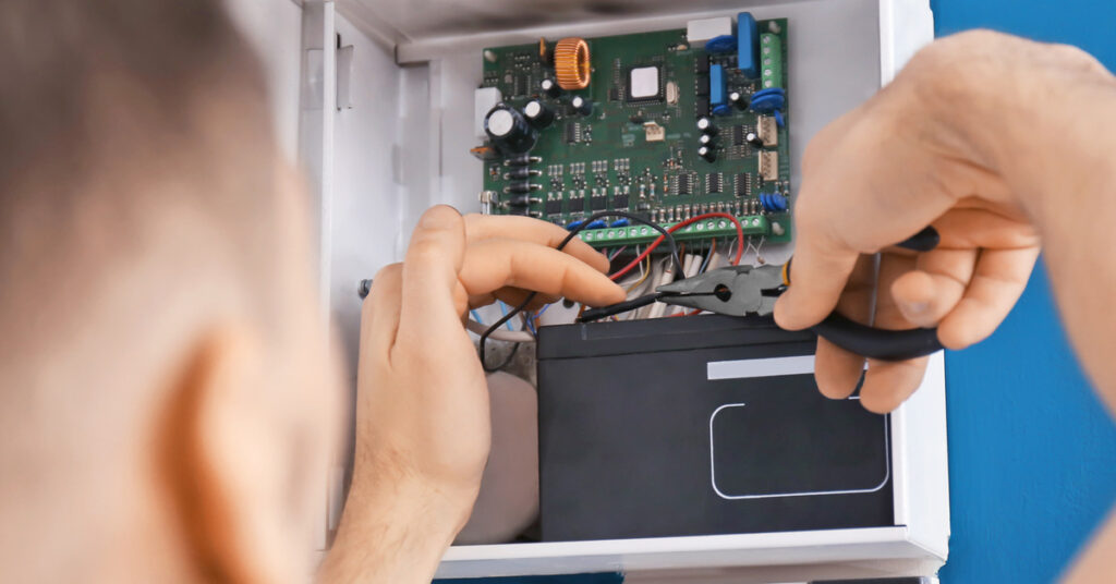 An open electrical box of an alarm system. There is a man working on the wires with a set of pliers.