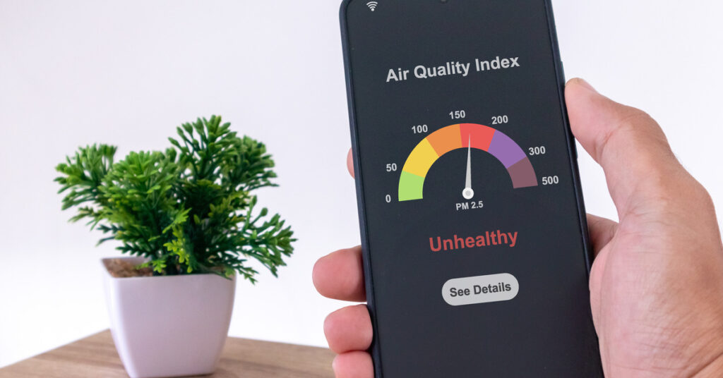 A person's hand is holding up a black smartphone in front of a fake plant. The screen displays an air quality index.